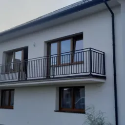 Nowoczesny, dwupiętrowy dom z białym tynkiem i czarną balustradą balkonową z pionowych prętów, widok z zewnątrz w słoneczny dzień.