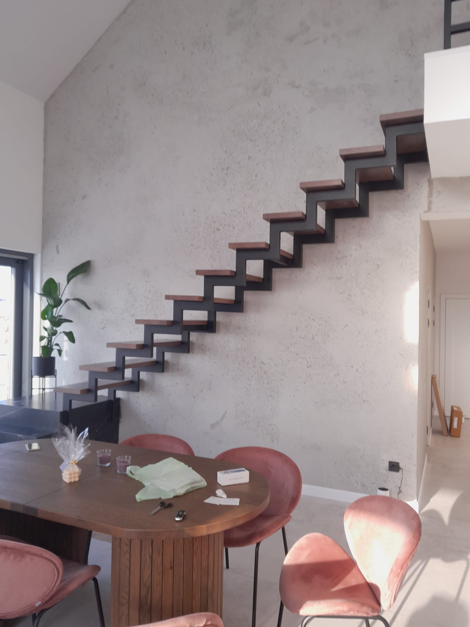Nowoczesne schody wspornikowe z drewnianymi stopniami i czarną metalową konstrukcją, zamontowane na szarej ścianie w minimalistycznym wnętrzu z widocznym stołem i krzesłami.