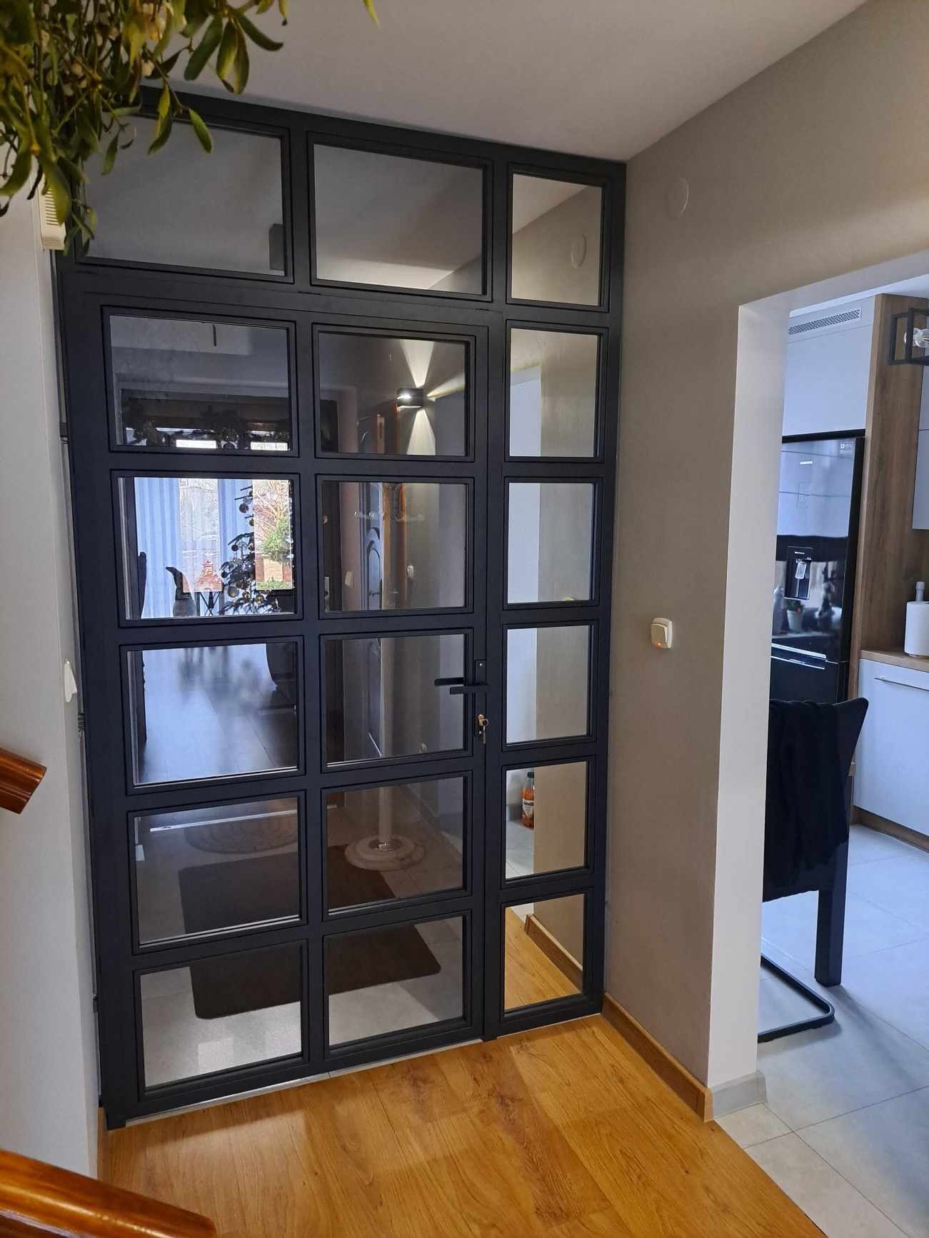 Szklana ściana działowa w metalowej ramie, z widocznym fragmentem schodów i wnętrzem domu, oświetlonym lampą sufitową.