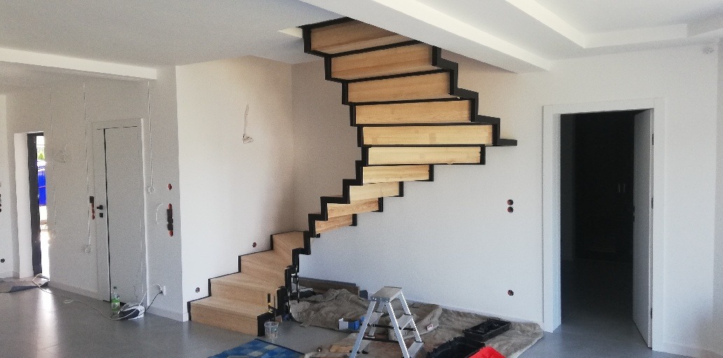 Nowoczesne, drewniane schody wspornikowe z czarną konstrukcją w jasnym wnętrzu, widoczna drabina i materiały zabezpieczające podłogę.