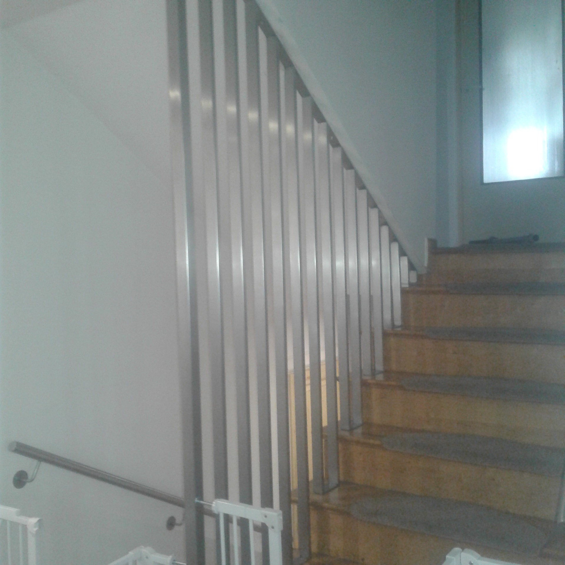 Metalowa balustrada schodowa z pionowymi szczeblami, widok z dołu schodów, obok drewniane schody z szarymi nakładkami.