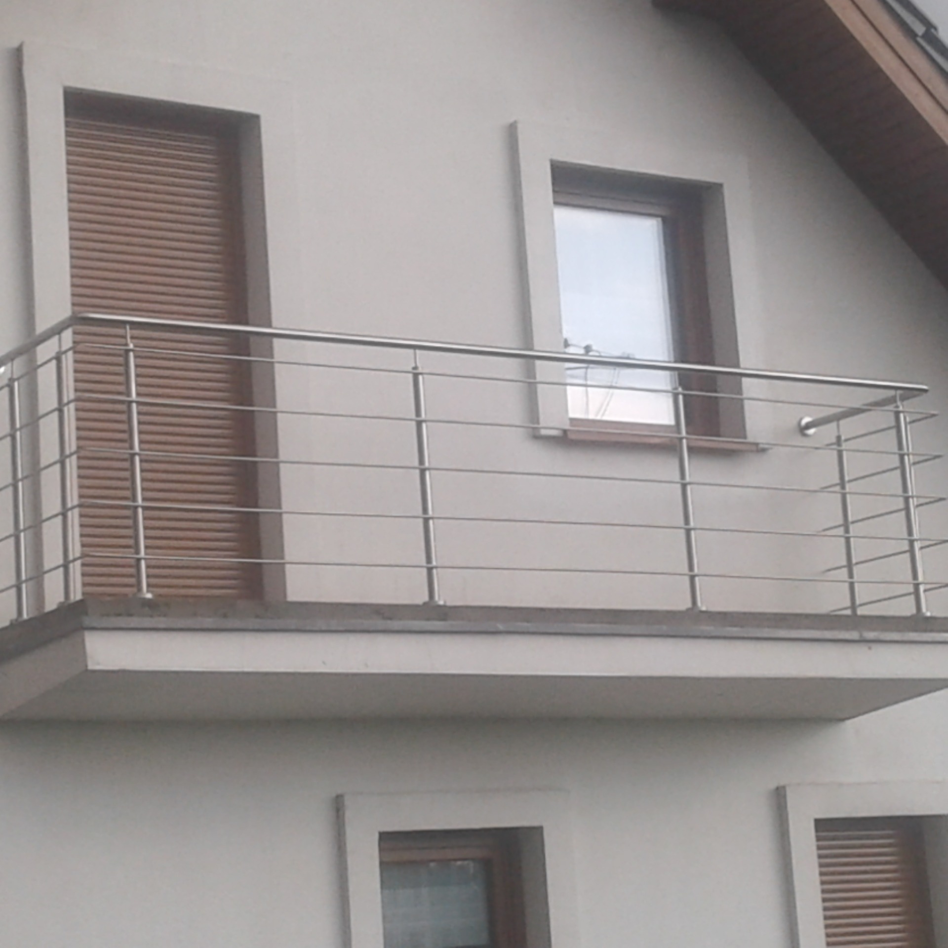 Biały budynek z balkonem z metalową balustradą, widoczne okna z brązowymi ramami i roletami.