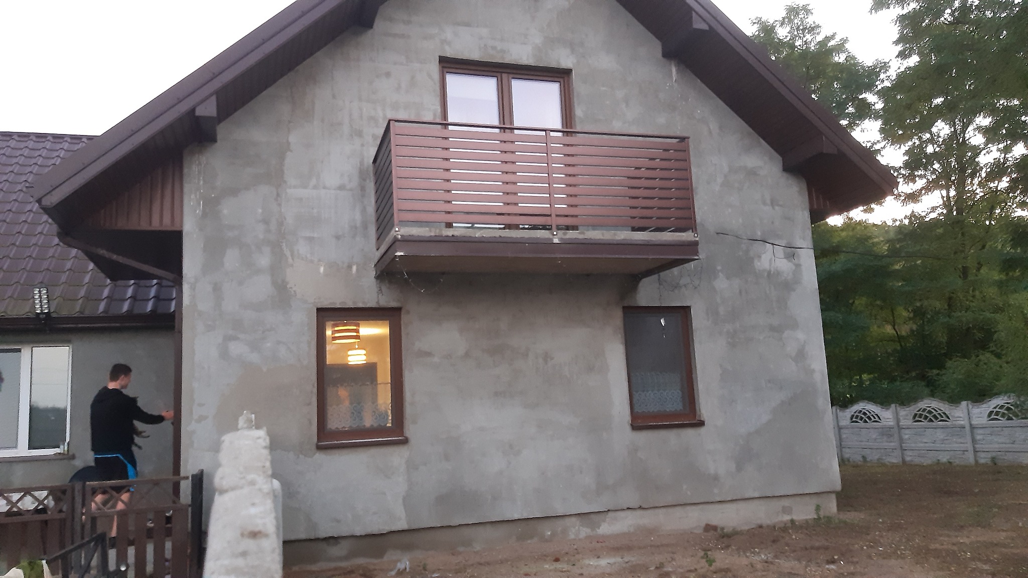 Szary dom z brązowym balkonem i oknami, widoczny mężczyzna przy ogrodzeniu, w tle zieleń.