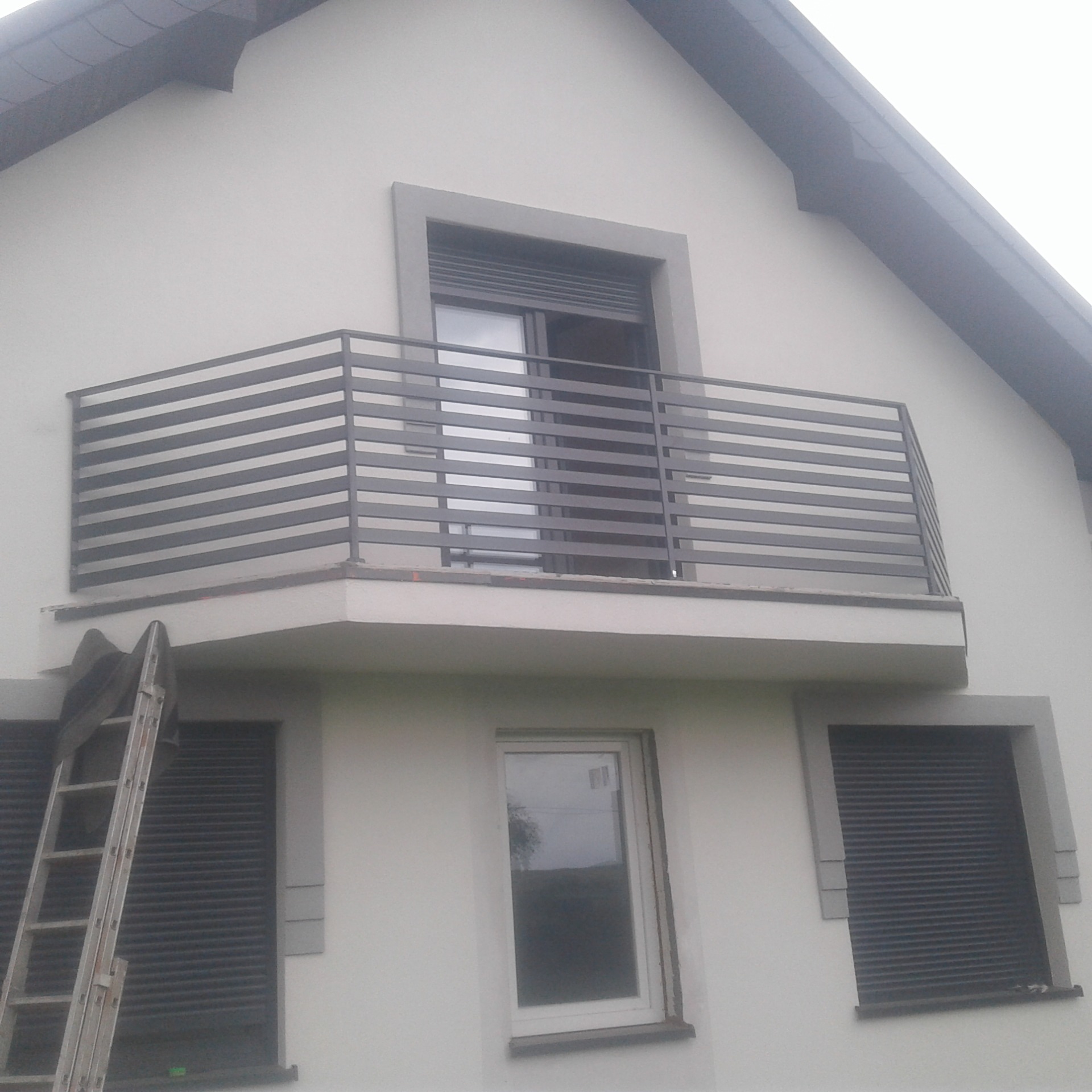 Nowoczesna, szara balustrada balkonowa z poziomych profili na tle białej fasady domu. Drewniana drabina oparta o ścianę obok okna z roletami.