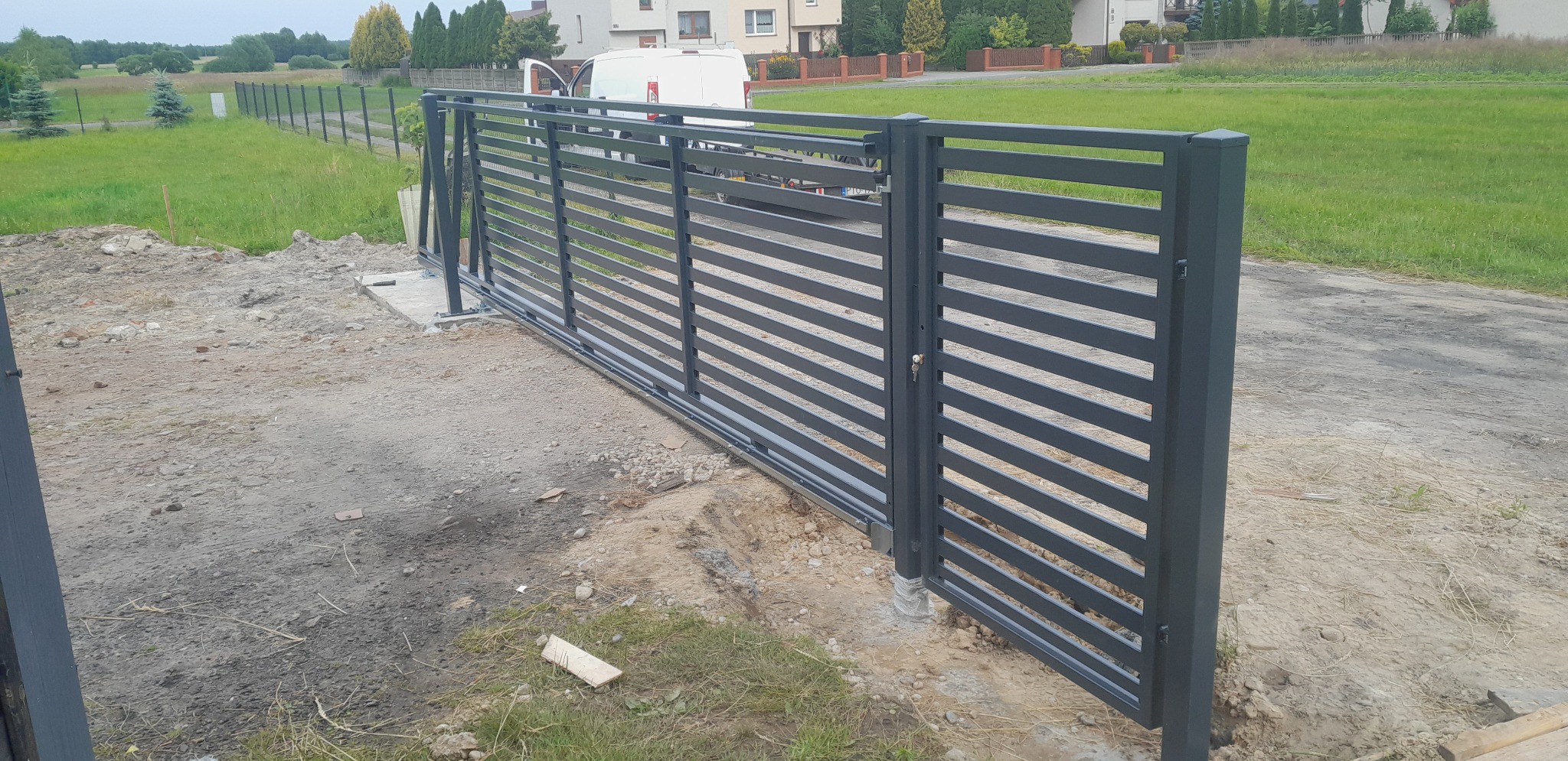 Nowoczesna, przesuwna brama wjazdowa z poziomymi panelami, pomalowana na grafitowo, zamontowana na betonowej podstawie, z widocznym mechanizmem przesuwnym i kluczem w zamku, na tle zieleni...