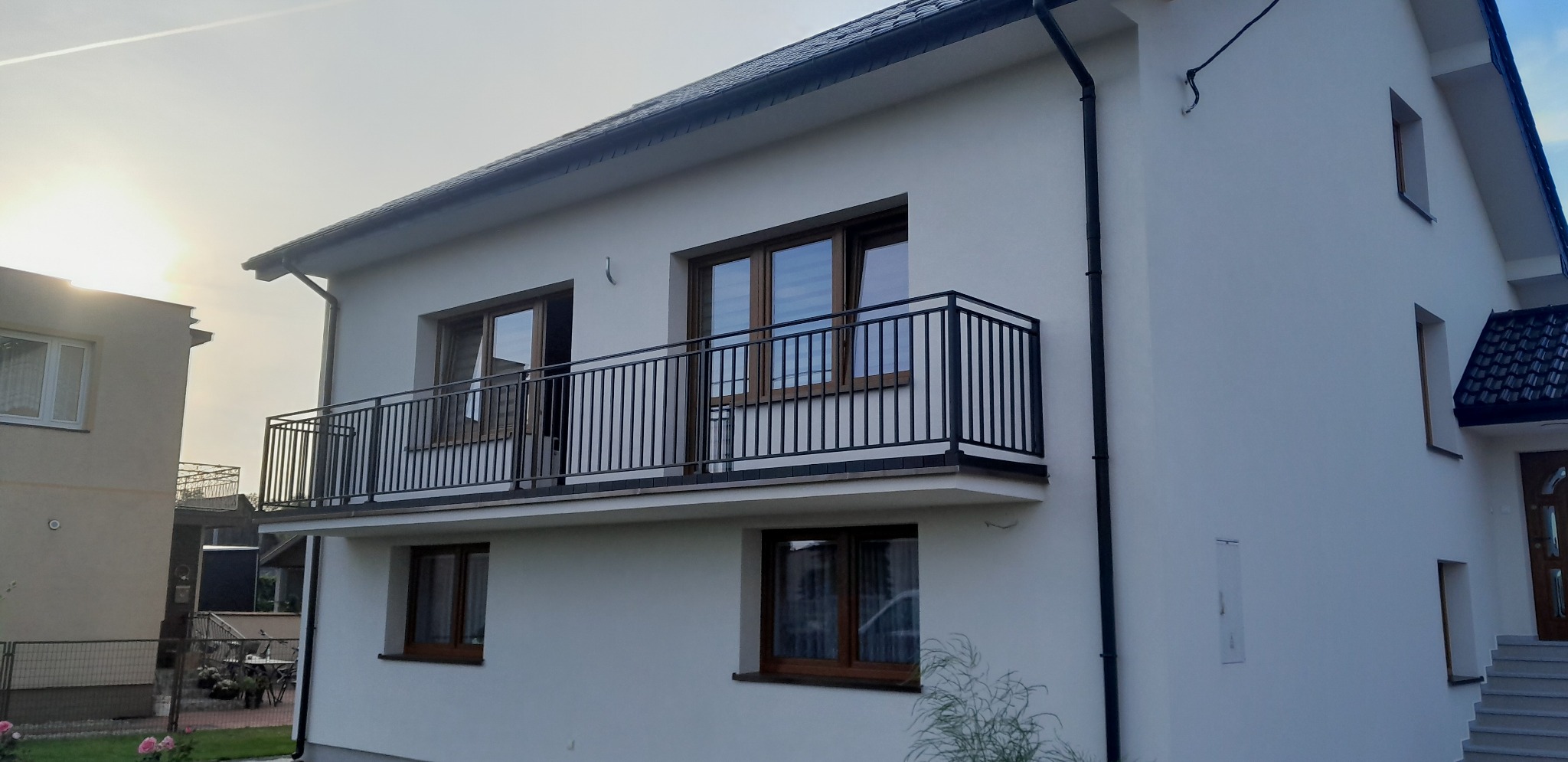 Nowoczesny, dwupiętrowy dom z białym tynkiem i czarną balustradą balkonową z pionowych prętów, widok z zewnątrz w słoneczny dzień.