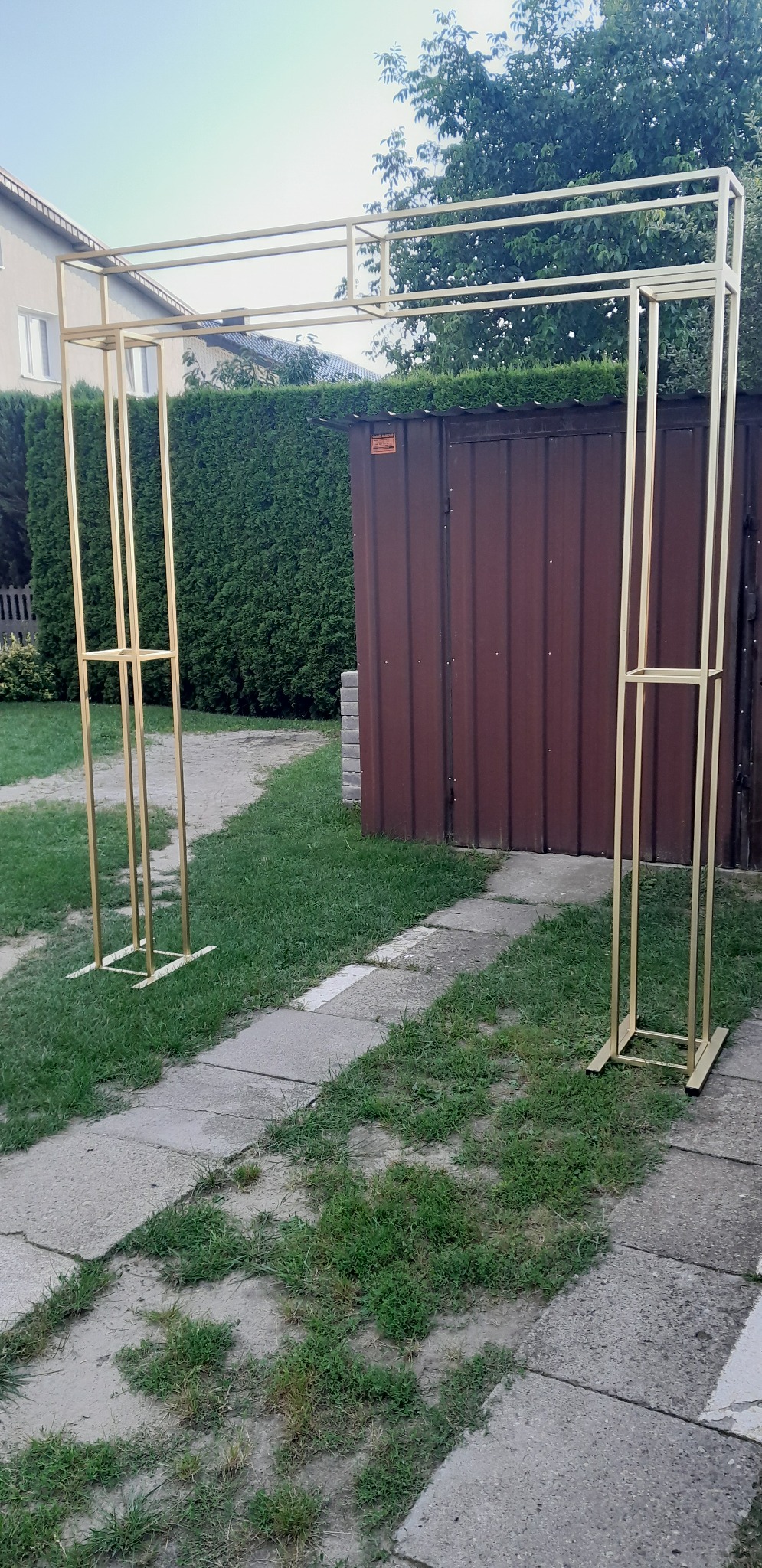 Metalowa konstrukcja w kształcie łuku, prawdopodobnie do dekoracji, ustawiona na trawniku przed brązową blaszana budką i żywopłotem.