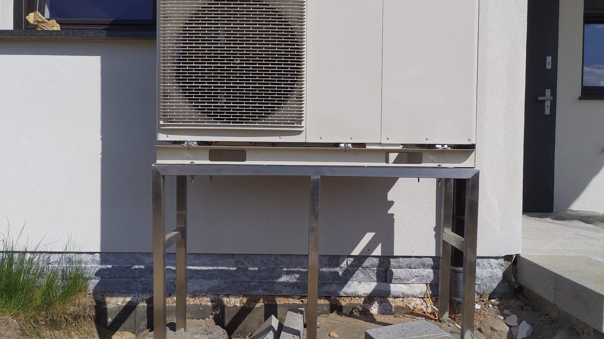 Zewnętrzna jednostka klimatyzacji zamontowana na metalowym stelażu przy ścianie budynku z widocznymi elementami instalacji i przygotowanym podłożem.