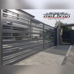 Nowoczesne ogrodzenie metalowe z poziomymi panelami w kolorze grafitowym, z logo firmy 'met.bron' widocznym w górnej części zdjęcia, na tle domu i kostki brukowej.