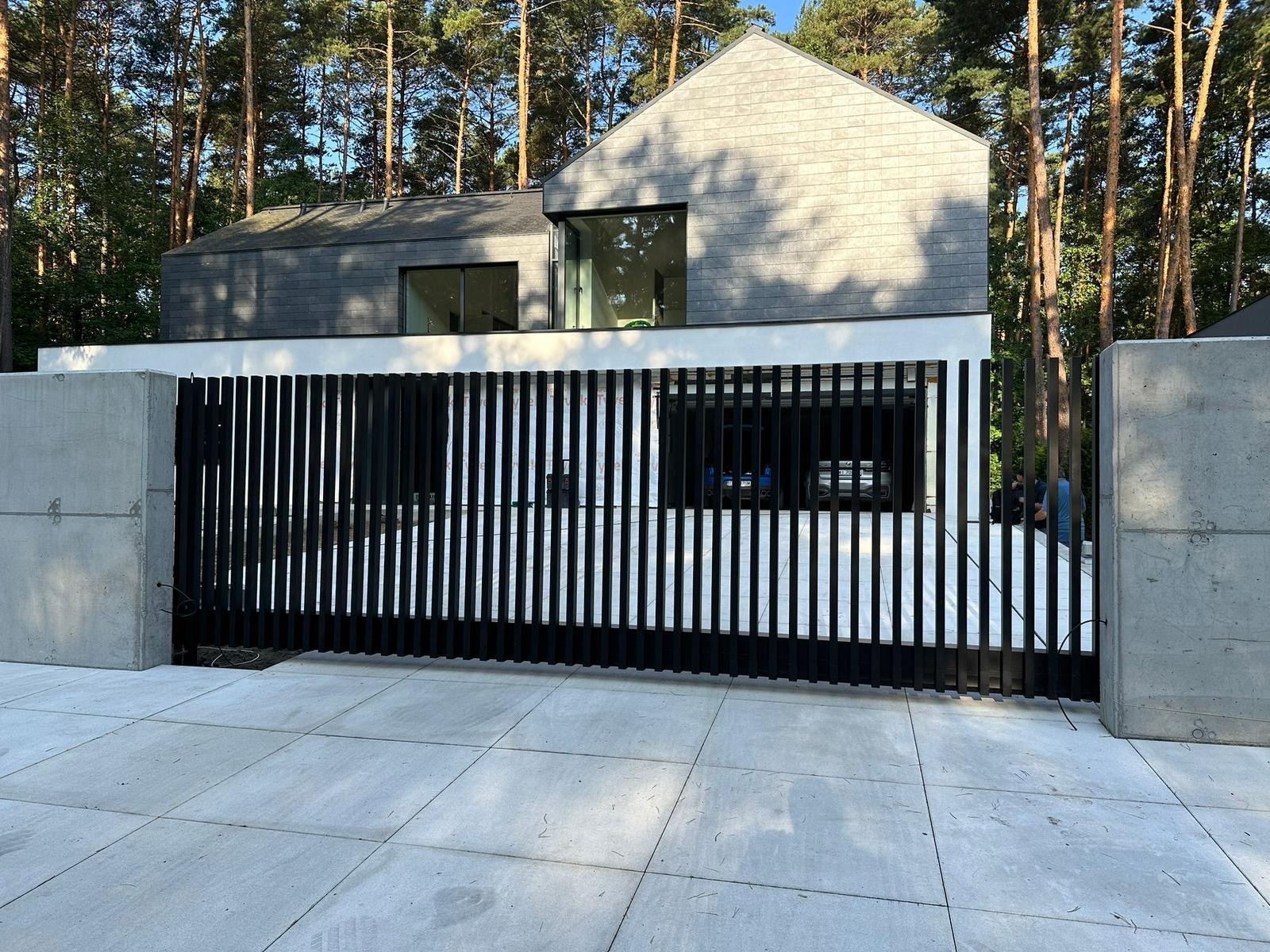 Nowoczesna posesja z przesuwną bramą z pionowych, czarnych paneli, osadzona między dwoma betonowymi słupkami, na tle domu z szarą dachówką i dużymi oknami.