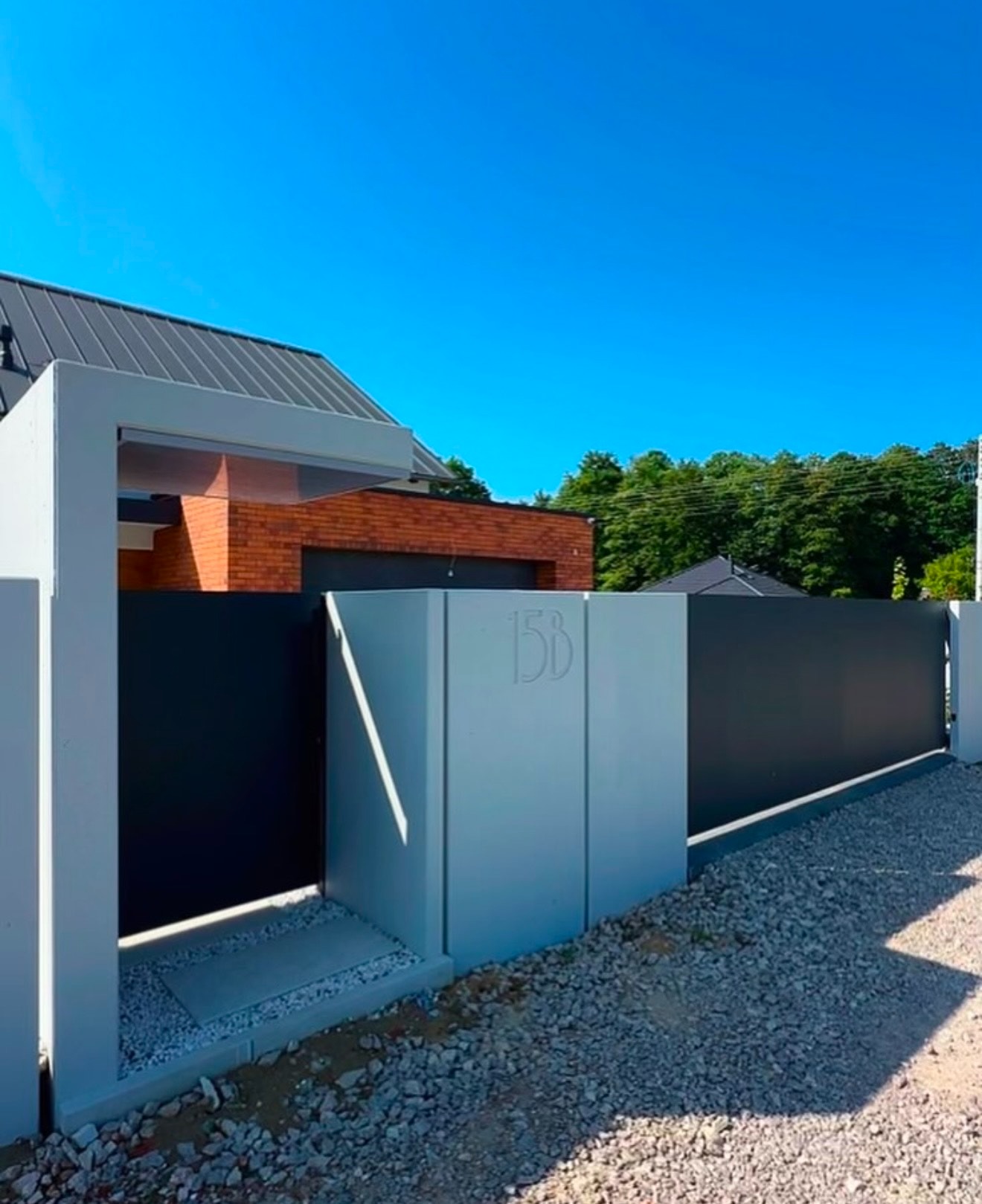 Nowoczesne ogrodzenie posesji z przesuwną bramą w kolorze antracytu, z betonowymi słupkami z numerem domu 15B, na tle domu z cegły i błękitnego nieba.