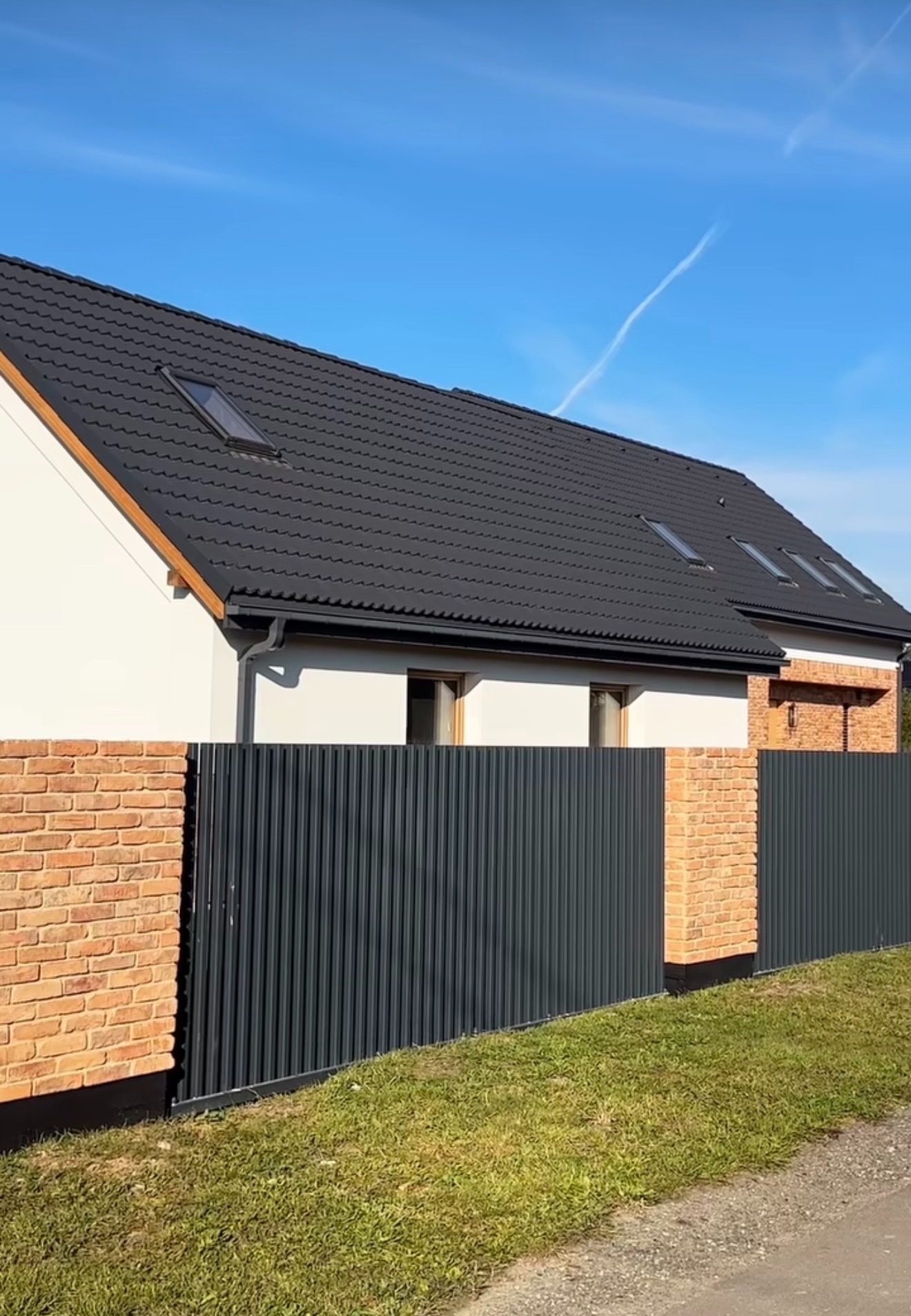 Nowoczesny dom jednorodzinny z ciemną, metalową elewacją ogrodzenia i ceglanymi słupkami, widok na dach z oknami dachowymi, błękitne niebo w tle.