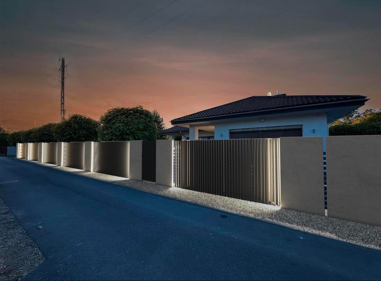 Nowoczesne ogrodzenie z pionowych paneli z wbudowanym oświetleniem LED, widok z poziomu ulicy o zmierzchu, w tle dom jednorodzinny.