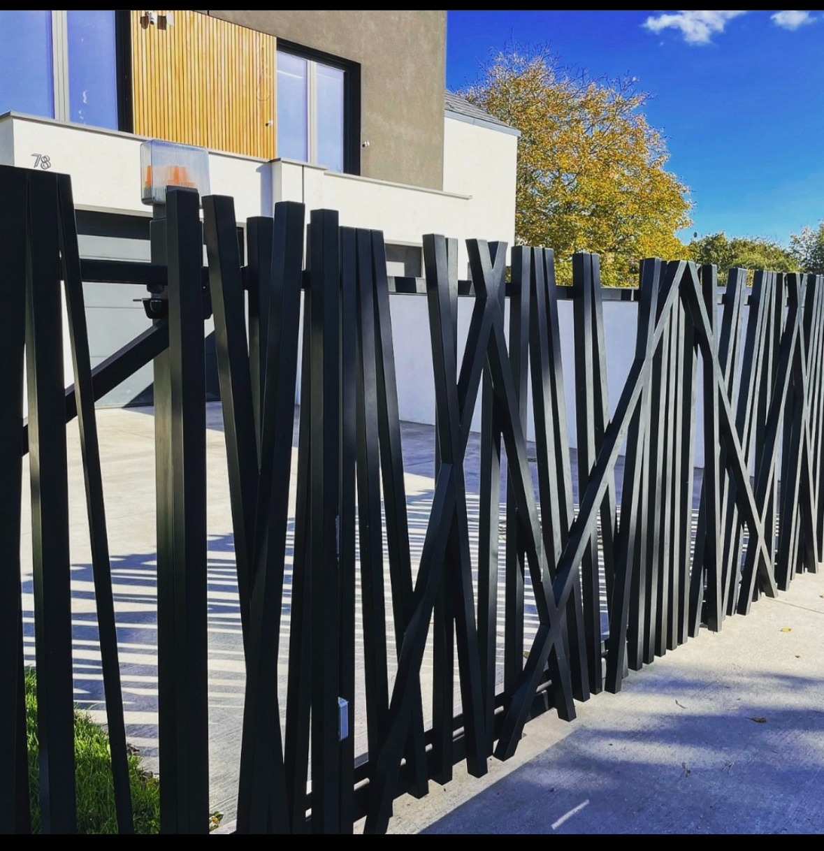 Nowoczesne ogrodzenie metalowe z czarnych, pionowych i ukośnych elementów, na tle domu z elementami drewna i błękitnego nieba.