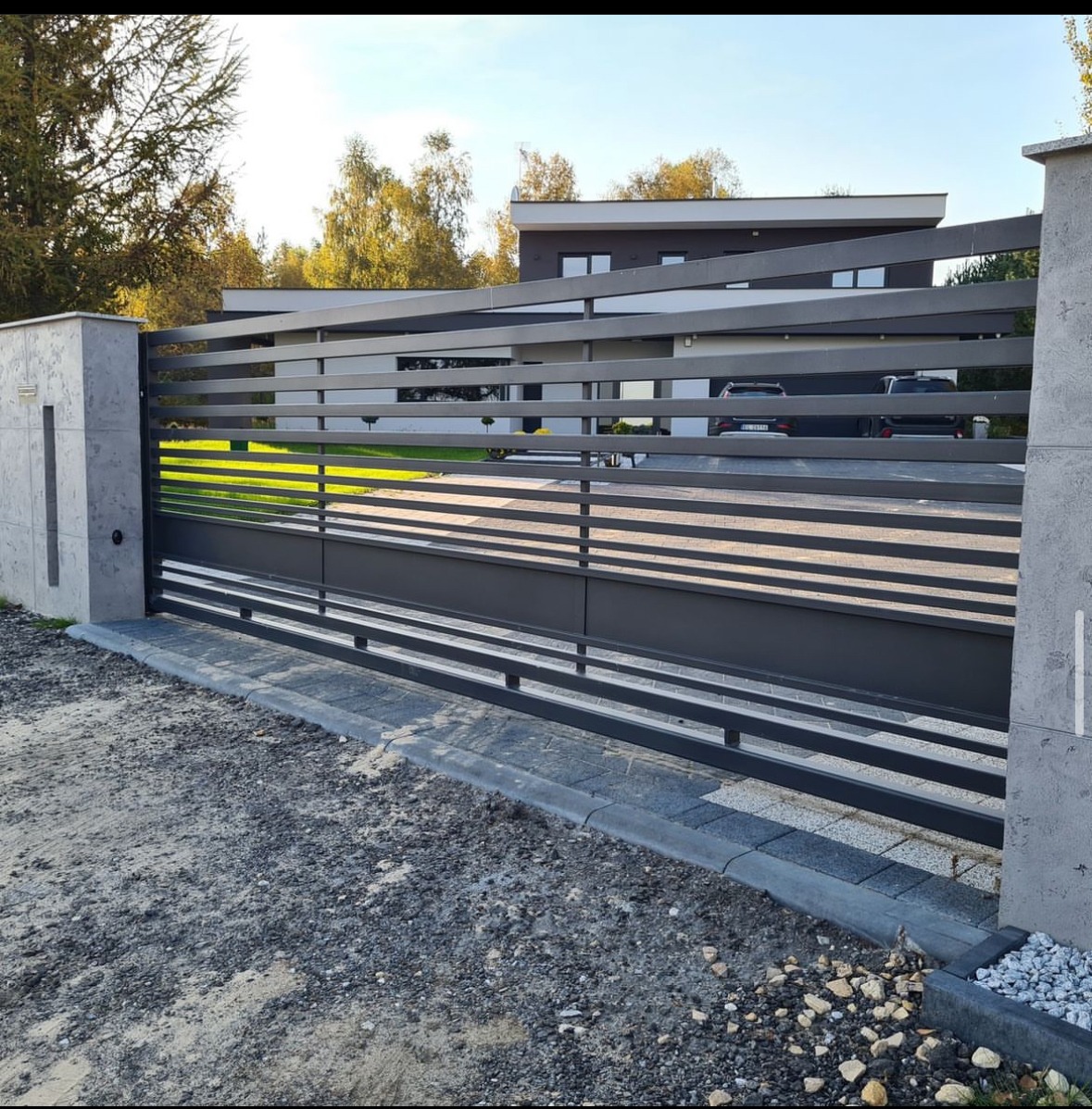 Nowoczesna, przesuwna brama wjazdowa z poziomych, metalowych profili, szara kolorystyka, na tle domu z płaskim dachem i zadbanym trawnikiem.