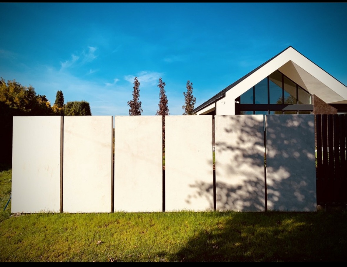 Nowoczesne, białe ogrodzenie z paneli na tle domu z dużymi oknami i błękitnego nieba, cień drzew na jednym z paneli.