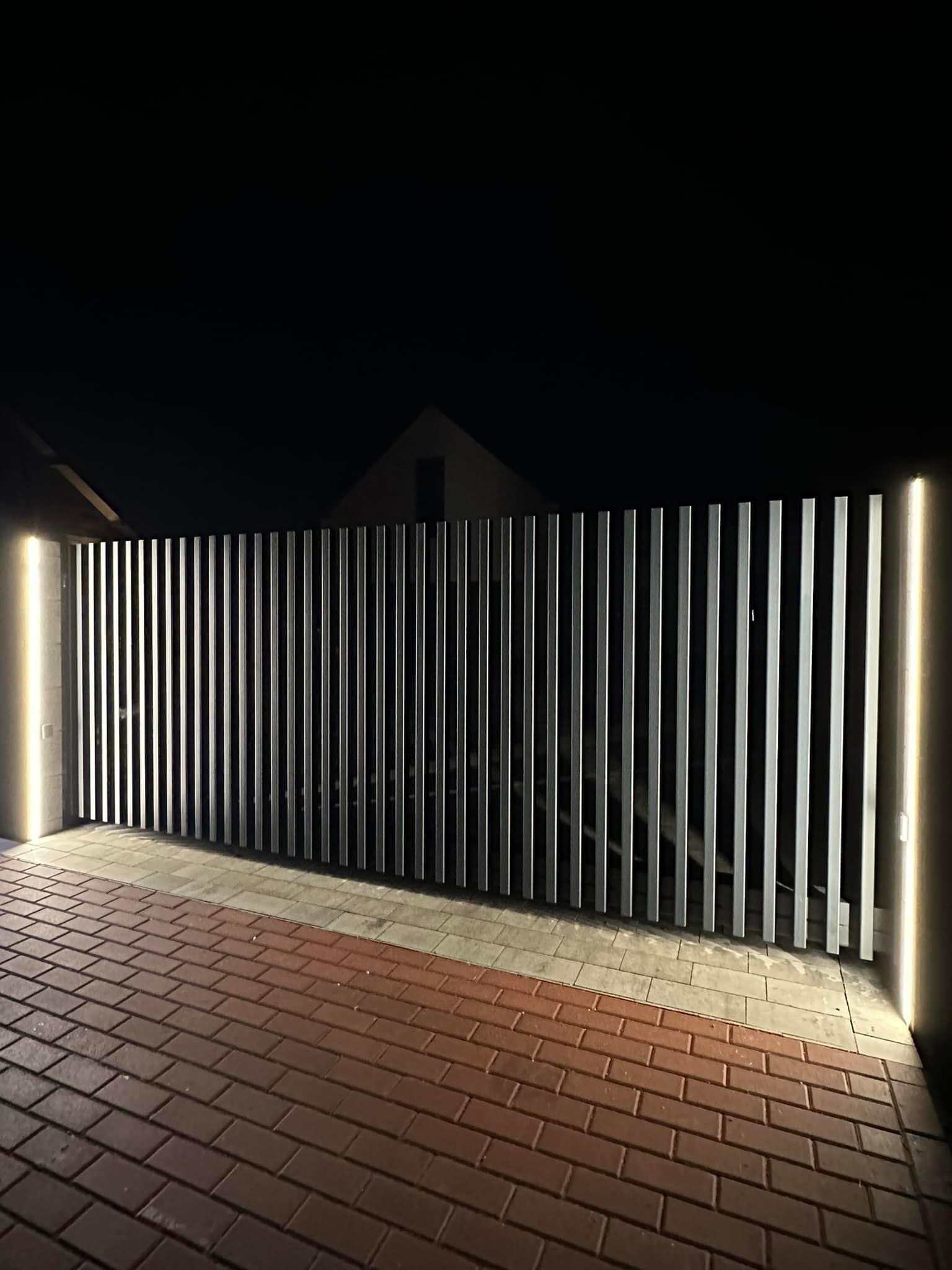Nowoczesne ogrodzenie z pionowych, metalowych paneli oświetlone pionowymi lampami po bokach, widoczne na tle ciemnego nieba i kostki brukowej.
