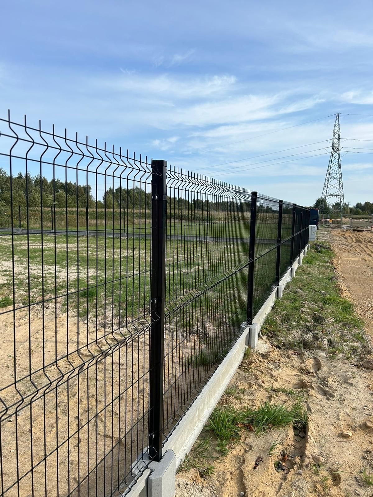 Długi, czarny system ogradzający z siatki, zamontowany na betonowej podmurówce, biegnący wzdłuż nierównego terenu z trawą i piaskiem, z widoczną w tle linią wysokiego napięcia.