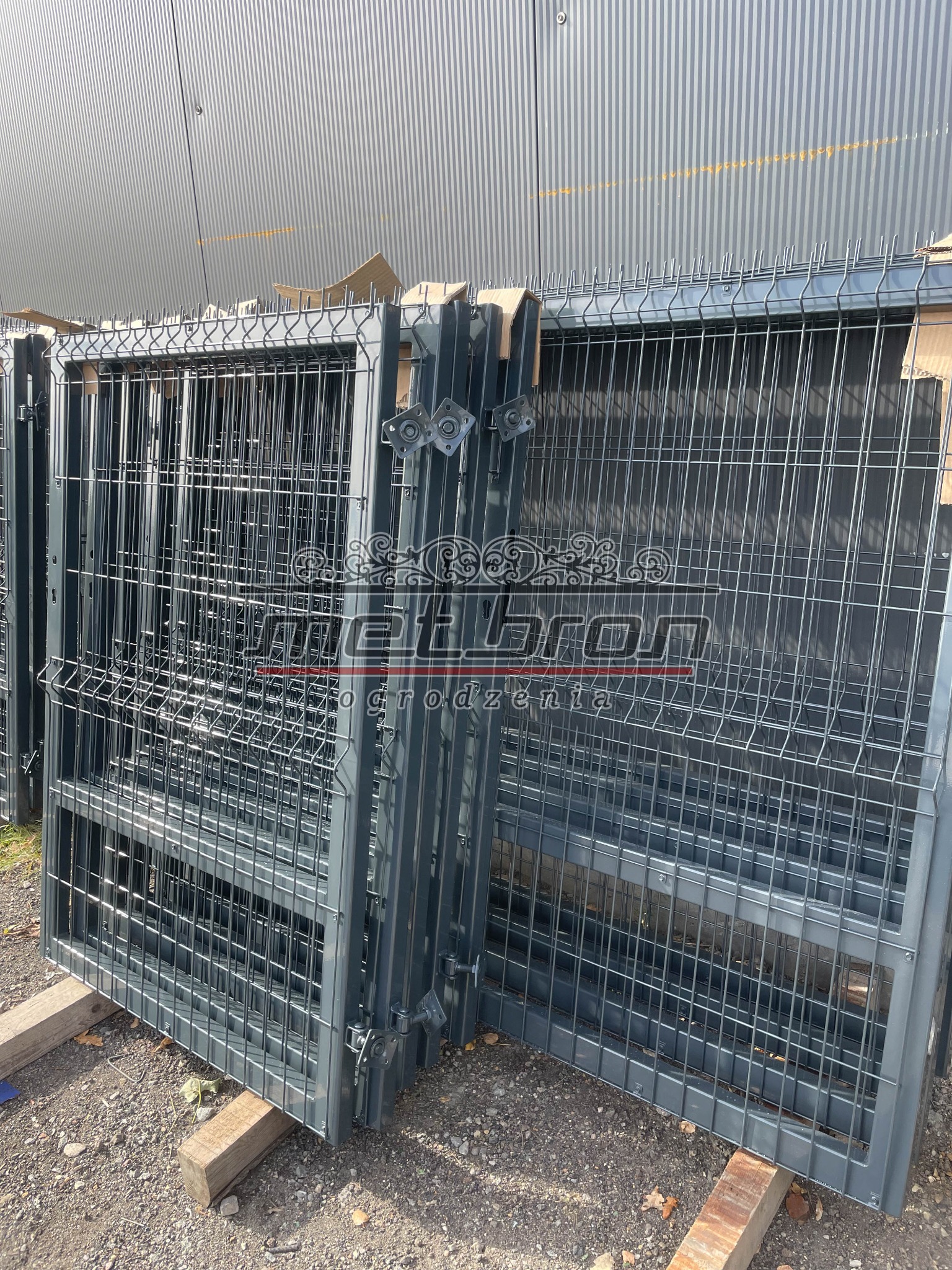 Stos paneli ogrodzeniowych 2D z drutu fi4mm, zabezpieczonych kartonem na górze, ułożonych na drewnianych belkach przed szarą, metalową ścianą magazynu.