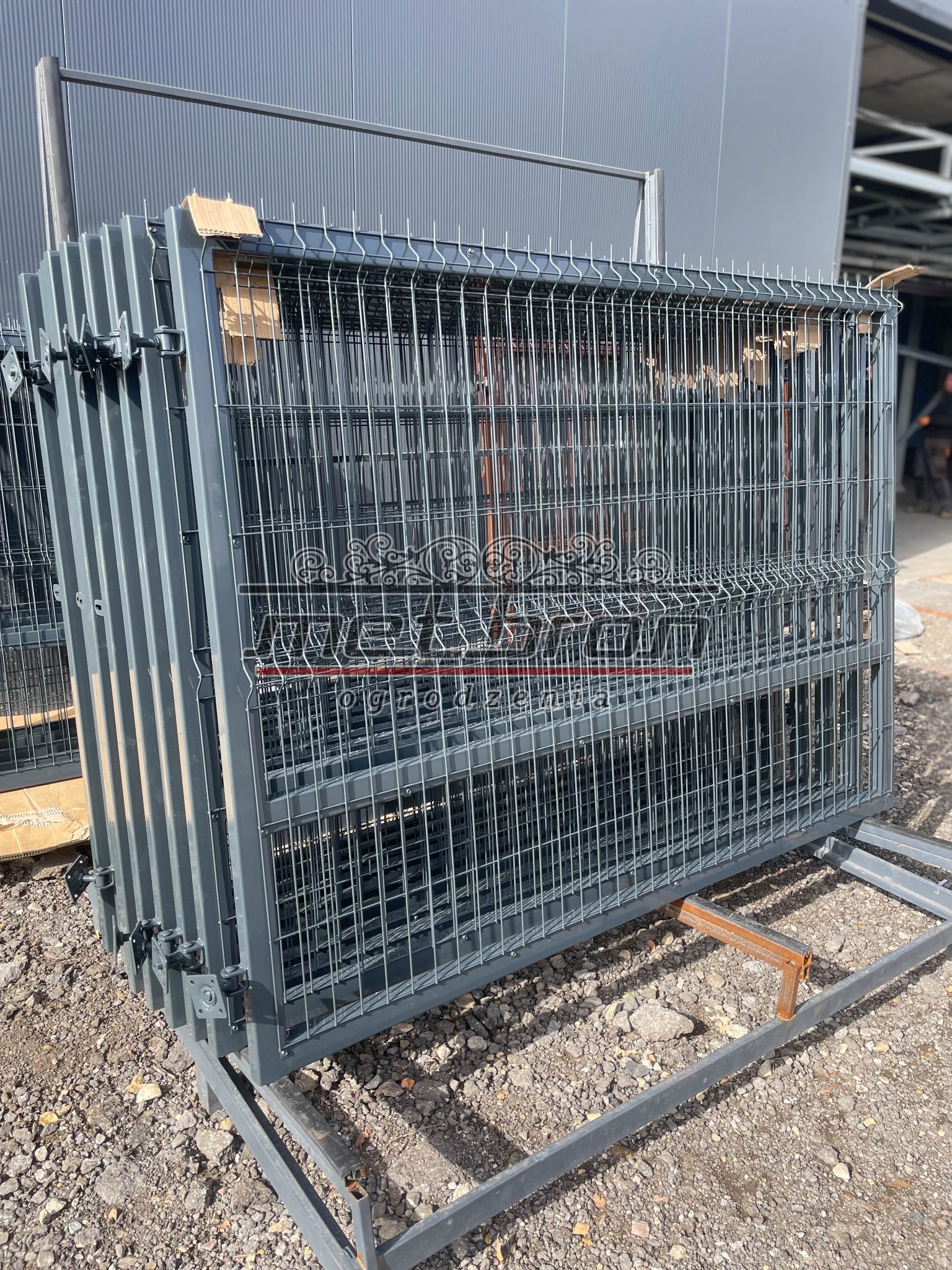 Stos paneli ogrodzeniowych z metalowej siatki, przygotowanych do transportu w Kłomnicach, widoczny na tle szarego budynku przemysłowego.