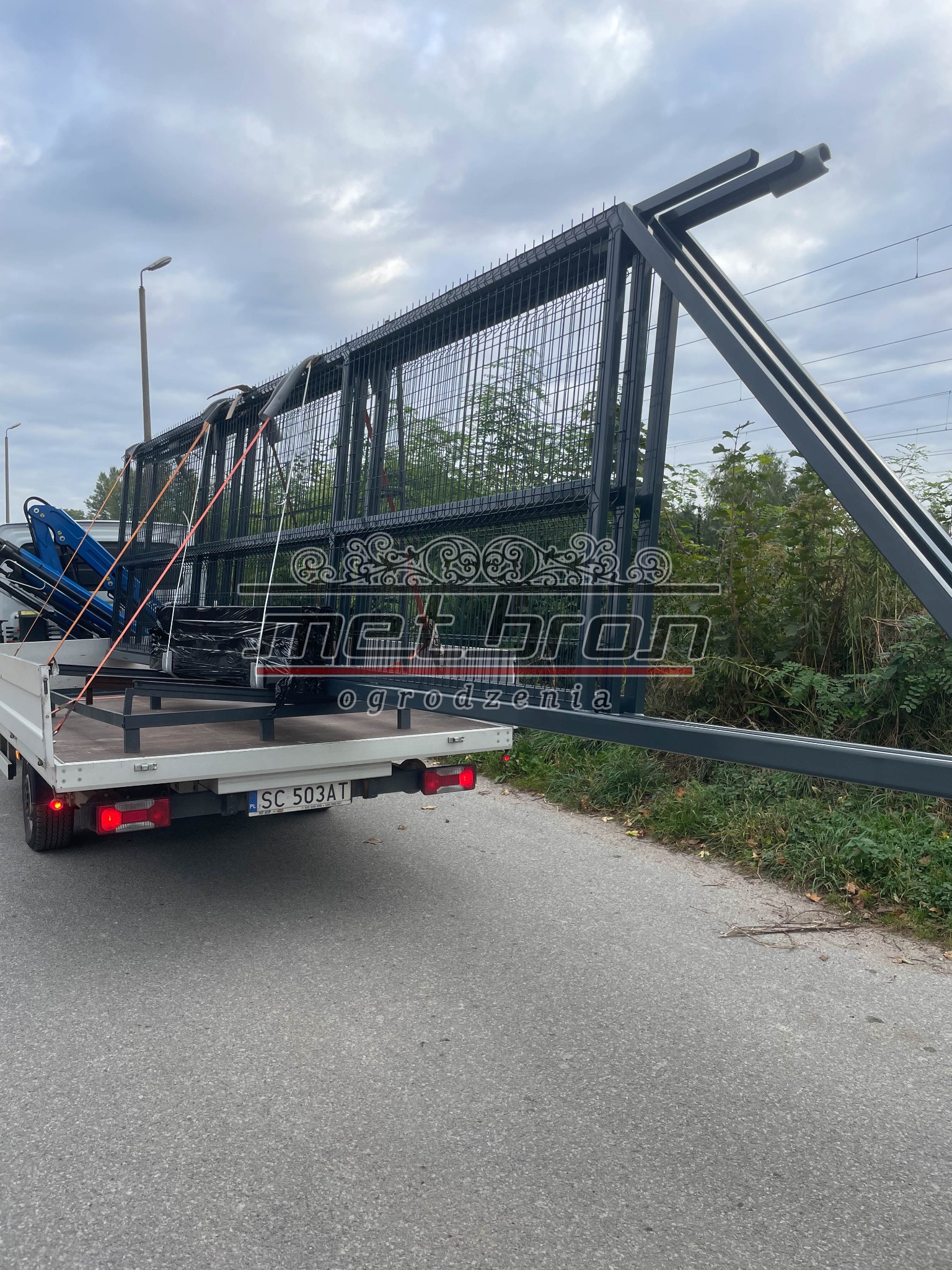 Transport ogrodzenia metalowego panelowego na przyczepie samochodu dostawczego, widoczne logo producenta, elementy montażowe i zabezpieczenia ładunku pasami.