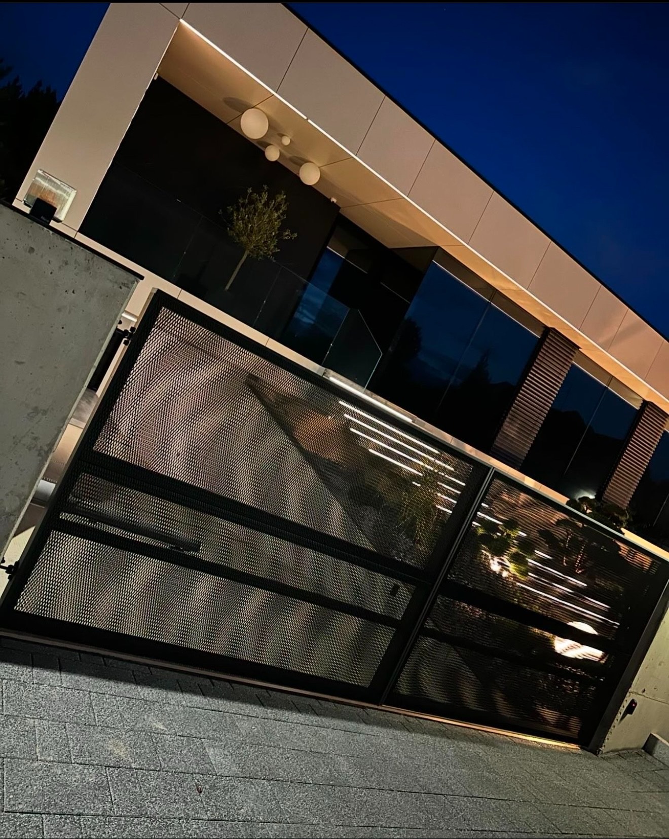 Nowoczesna, czarna, metalowa brama z perforowanymi panelami na tle domu z oświetleniem zewnętrznym i szklanymi elementami, widok z dołu.