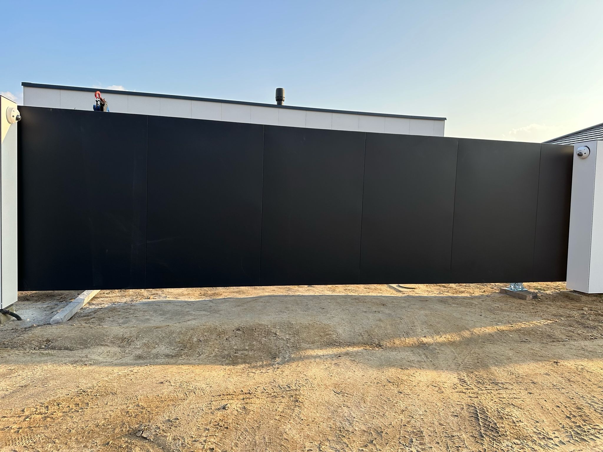 Nowoczesna, przesuwna brama panelowa w kolorze antracytowym, z widocznymi elementami montażowymi na gruncie i kamerą monitoringu na słupku, na tle budynku z jasną elewacją.