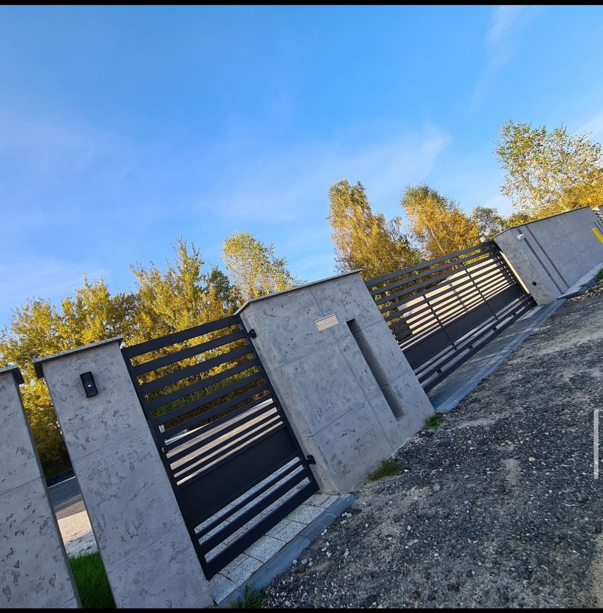 Nowoczesne ogrodzenie z poziomych, czarnych profili metalowych, osadzone w szarych, betonowych słupkach z wnęką na skrzynkę pocztową, widok pod kątem, błękitne niebo w tle.