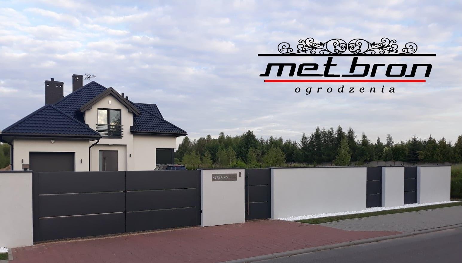 Nowoczesne ogrodzenie posesji z przesuwną bramą w kolorze antracytowym, białe słupki, widoczny dom z ciemnym dachem i logo firmy nad ogrodzeniem.