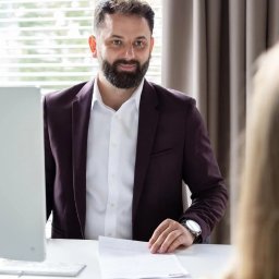 Kredytowy Poradnik sp. z o.o. - Uśmiechnięty doradca w bordowej marynarce siedzi za biurkiem z białym blatem, za nim okno z żaluzjami i zasłony, na biurku leżą dokumenty i stoi monitor komputera.