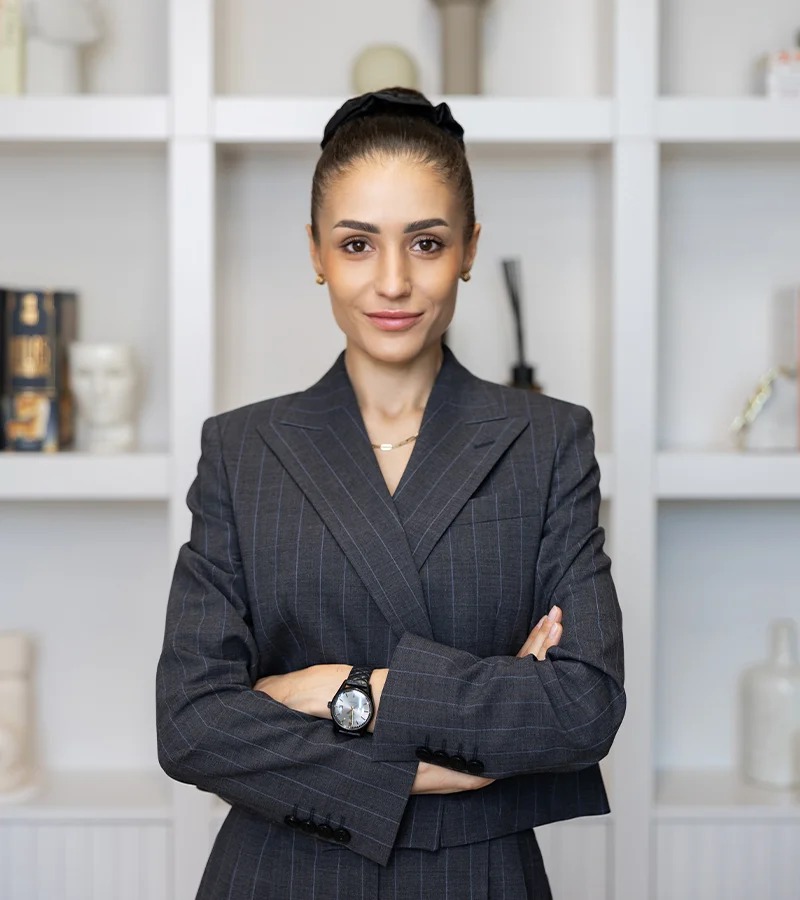 Portret kobiety w szarym, prążkowanym garniturze, ze skrzyżowanymi rękami i zegarkiem na ręce, stojącej na tle białego regału z dekoracjami.