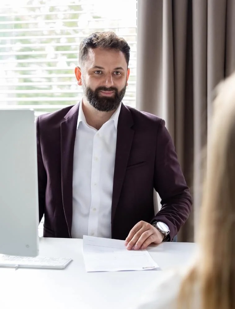 Uśmiechnięty doradca w bordowej marynarce siedzi za biurkiem z białym blatem, za nim okno z żaluzjami i zasłony, na biurku leżą dokumenty i stoi monitor komputera.