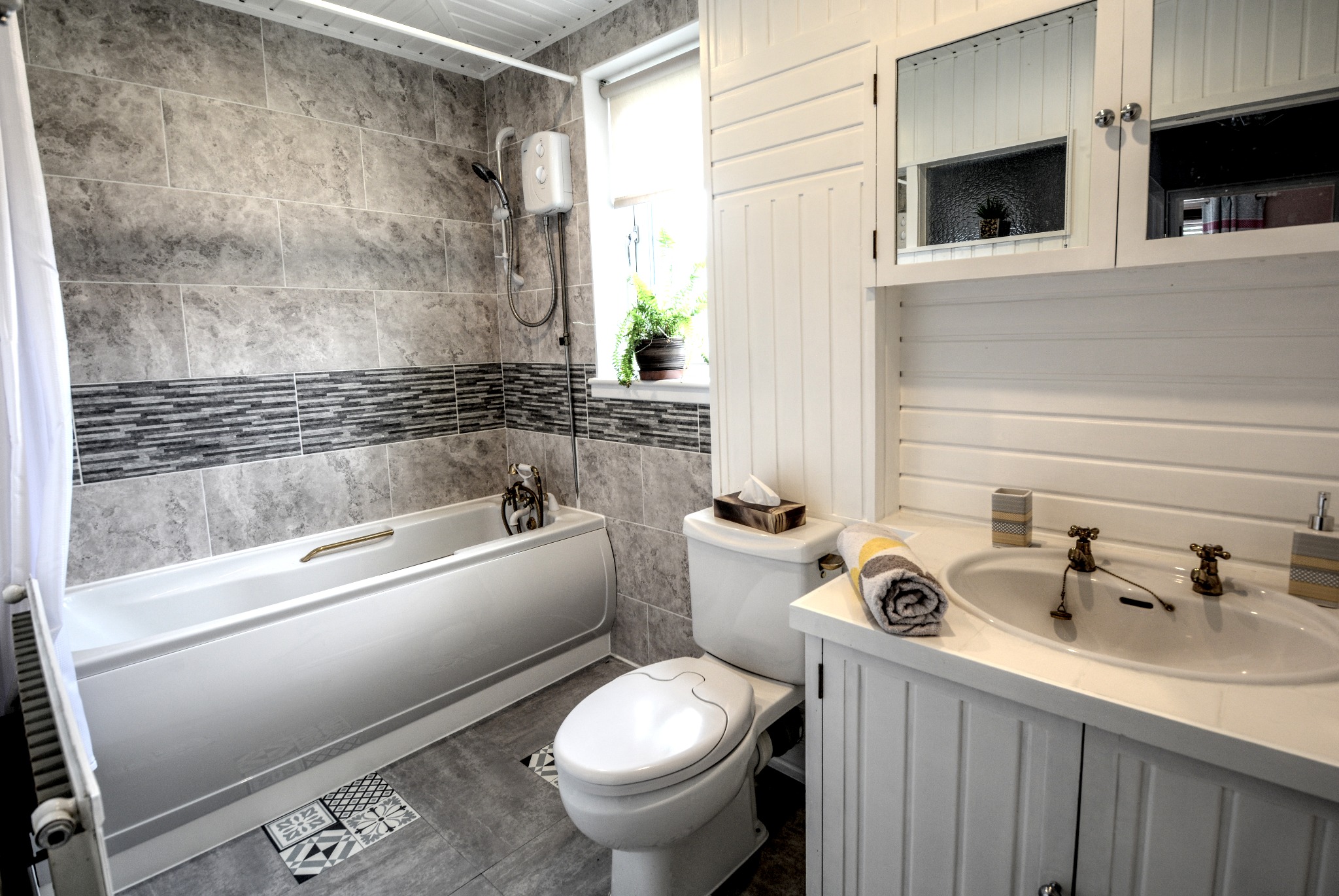 Wykończona łazienka w szaro-białej tonacji z wanną, toaletą i umywalką. Na ścianach szare płytki z dekoracyjnym pasem. Roślina w oknie dodaje naturalnego akcentu.
