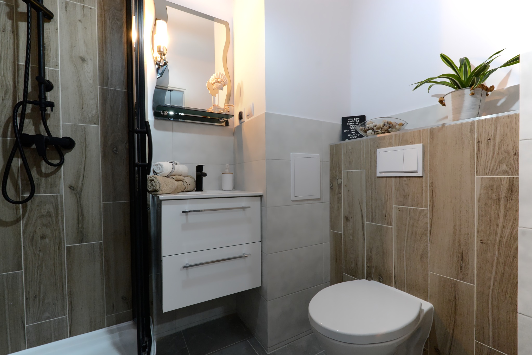 Nowoczesna łazienka z kabiną prysznicową typu walk-in, drewnopodobnymi płytkami i białymi meblami. Uwagę zwraca czarna armatura i dekoracje.