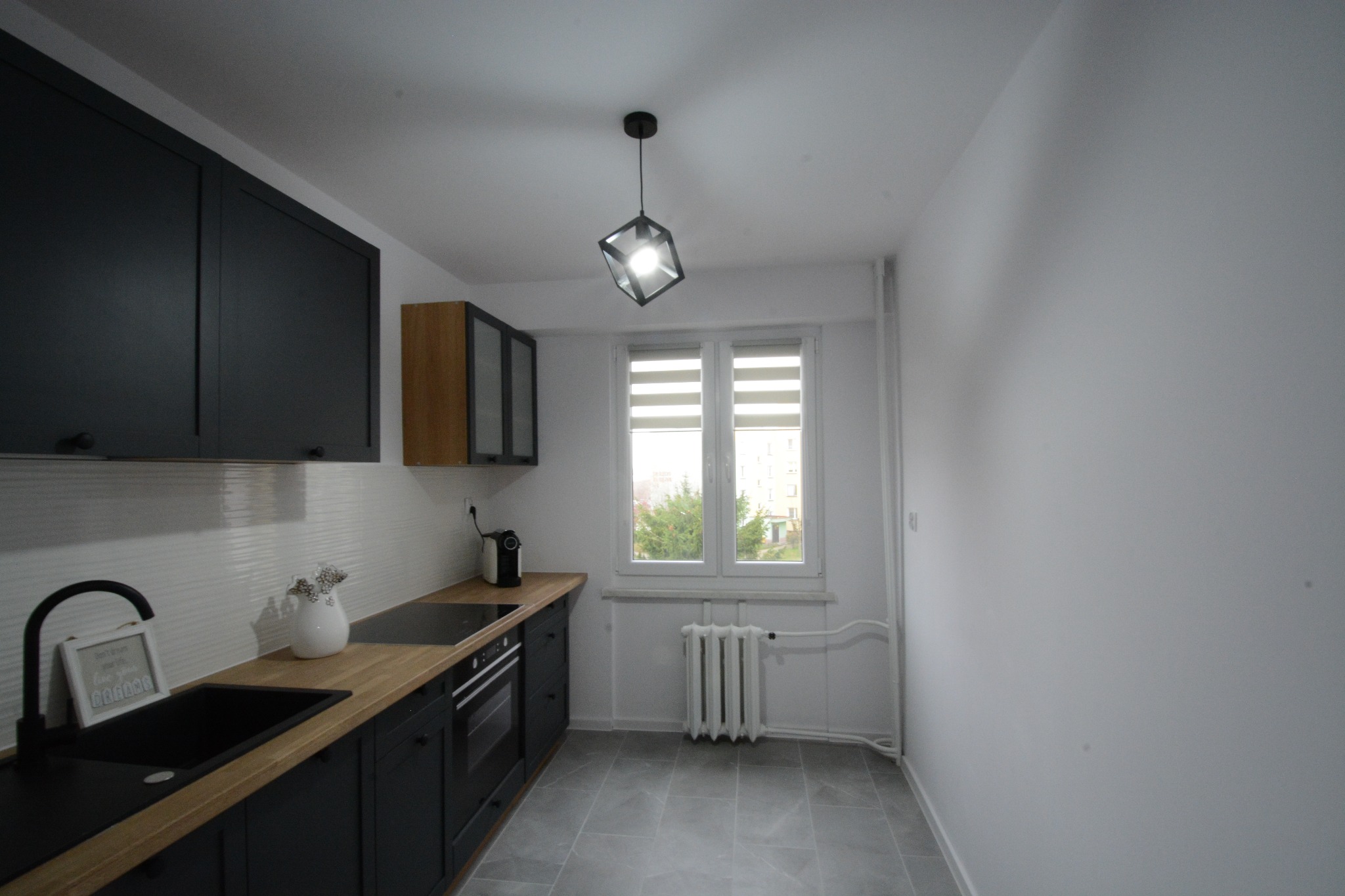 Nowoczesna kuchnia z czarnymi szafkami, drewnianym blatem i szarą podłogą. Geometryczna lampa wisząca nad oknem. Minimalistyczny design z akcentem na funkcjonalność.