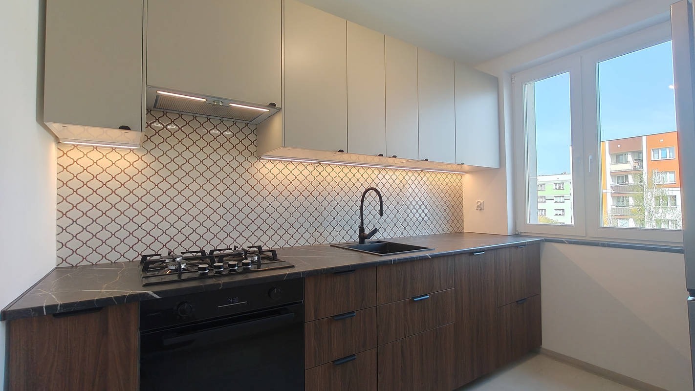 Nowoczesna kuchnia z beżowymi szafkami górnymi, drewnopodobnymi dolnymi i mozaiką nad blatem. Widok na okno z widokiem na budynek mieszkalny.