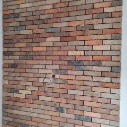 WTL-BUD - Ściana wyłożona dekoracyjną cegłą w odcieniach brązu, czerwieni i szarości, z widocznym gniazdkiem elektrycznym i wystającymi przewodami, na tle surowej, nie wykończonej podłogi.