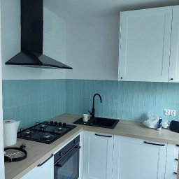 WTL-BUD - Nowoczesna, jasna kuchnia w Poznaniu z turkusowymi płytkami na ścianie, białymi szafkami z drewnianym blatem, czarnym zlewem i płytą grzewczą oraz widocznym wiaderkiem farby na podłodze.