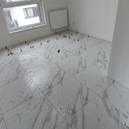 WTL-BUD - Układanie białych płytek podłogowych z szarym marmurkowym wzorem, z użyciem poziomujących klipsów i klinów w nowym, jasnym pomieszczeniu z oknem.