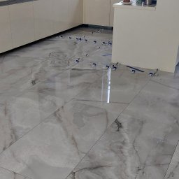 WTL-BUD - Błyszczące, szare płytki podłogowe z wzorem marmuru, ułożone w kuchni, z widocznymi niebieskimi klinami poziomującymi i odblaskiem okna.