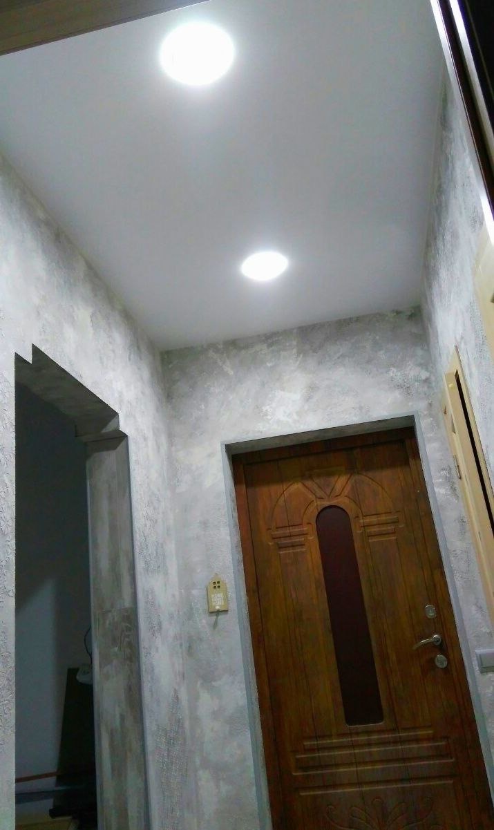 Wnętrze korytarza z ozdobnymi ścianami imitującymi beton, drewnianymi drzwiami z szybą i okrągłymi lampami sufitowymi.