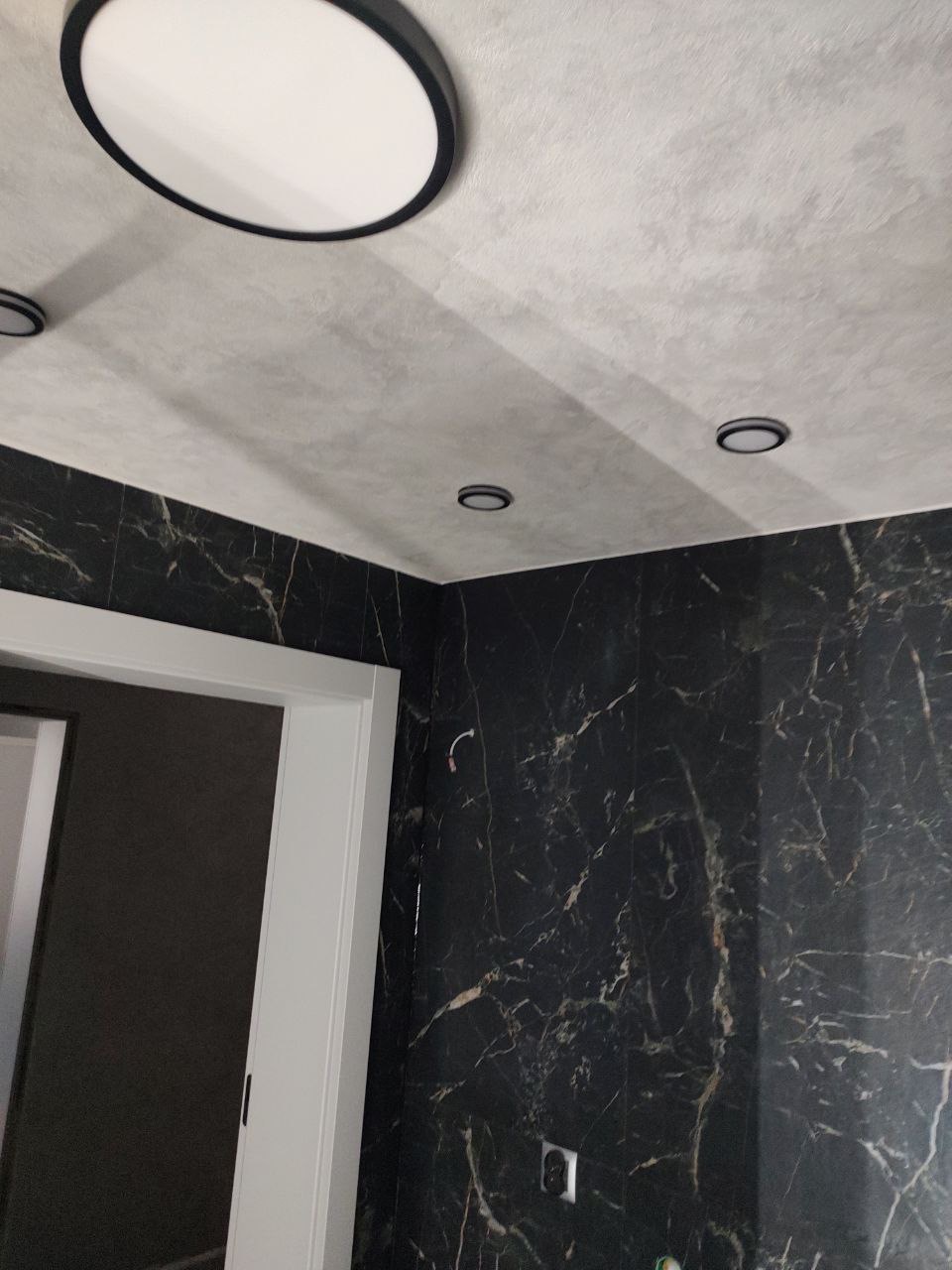 Wykończona łazienka z czarnymi płytkami imitującymi marmur na ścianach, szarą gładzią na suficie i nowoczesnym oświetleniem punktowym oraz okrągłą lampą sufitową.