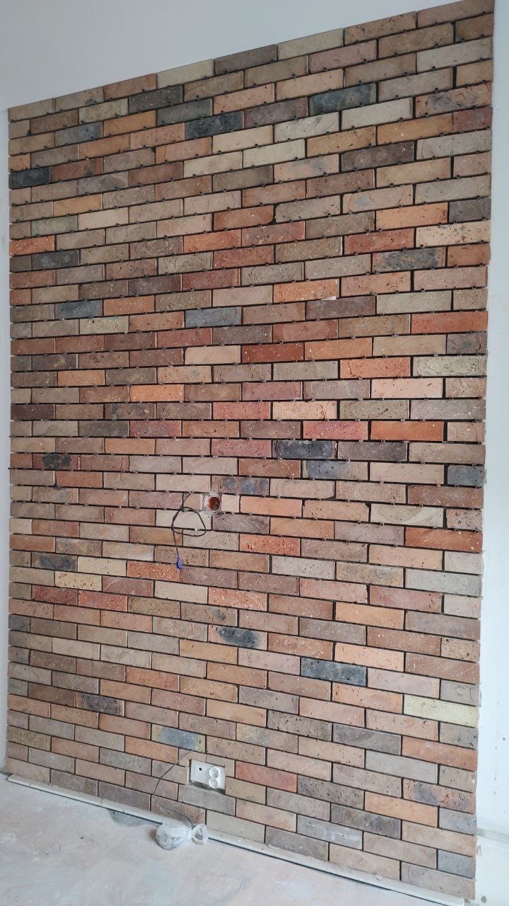 Ściana wyłożona dekoracyjną cegłą w odcieniach brązu, czerwieni i szarości, z widocznym gniazdkiem elektrycznym i wystającymi przewodami, na tle surowej, nie wykończonej podłogi.