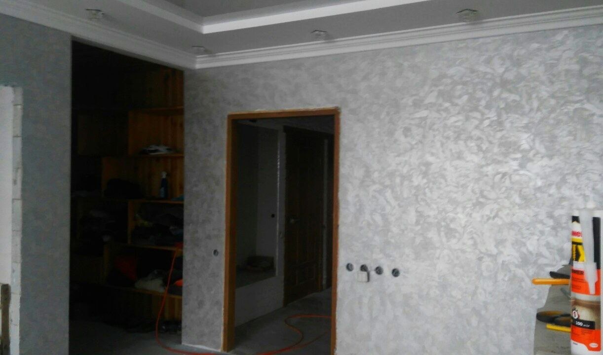 Ściana pokryta dekoracyjną, srebrzystą fakturą z motywem liści, widoczne puszki z silikonem i narzędzia, fragment ościeżnicy drzwiowej i otwarta szafa w tle.