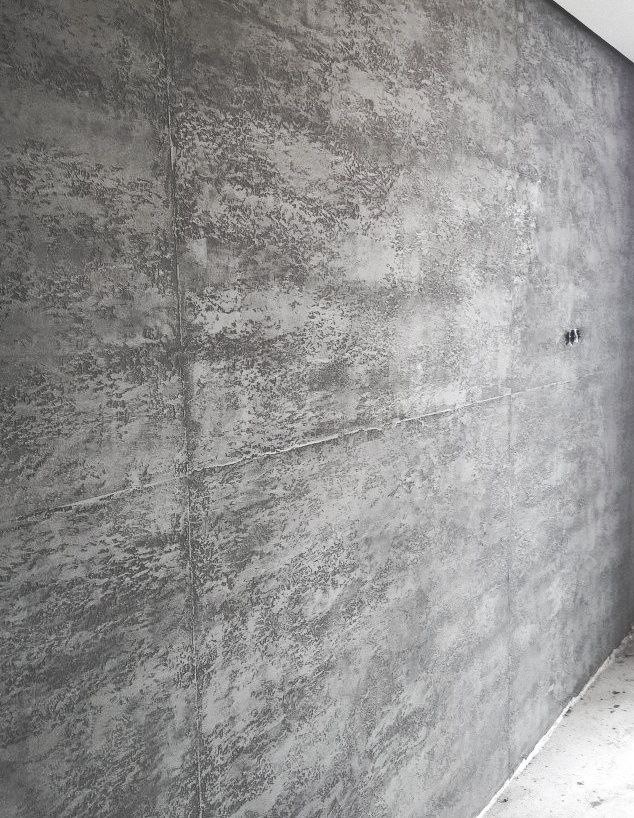 Szara ściana z efektem dekoracyjnym, imitującym beton architektoniczny, z widocznymi śladami po szpachli i delikatną fakturą, małe gniazdko elektryczne po prawej stronie.