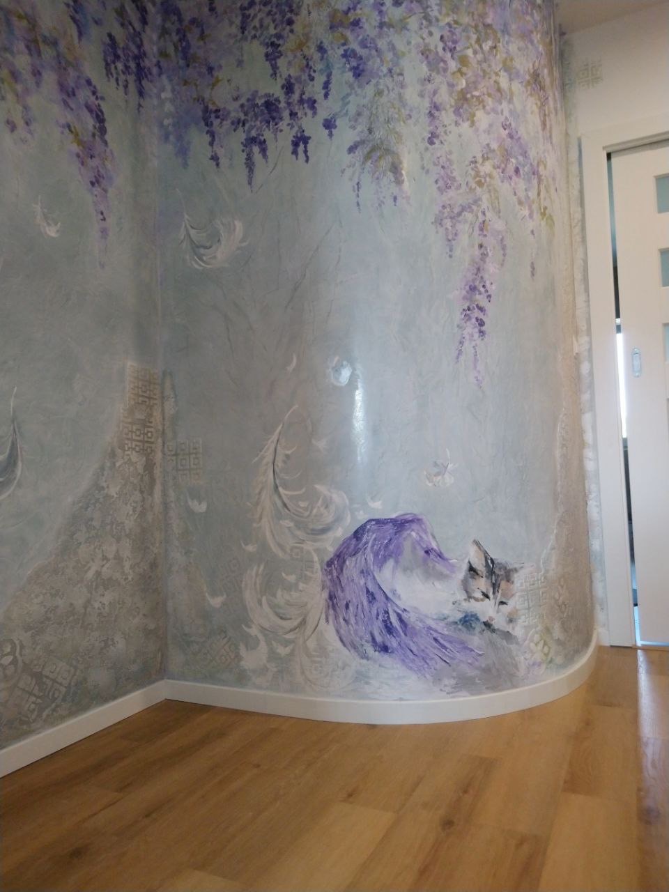 Ściana pokryta dekoracyjną gładzią z motywem wisterii i śpiącego kota, wykończona listwą przypodłogową, z widoczną podłogą z paneli.