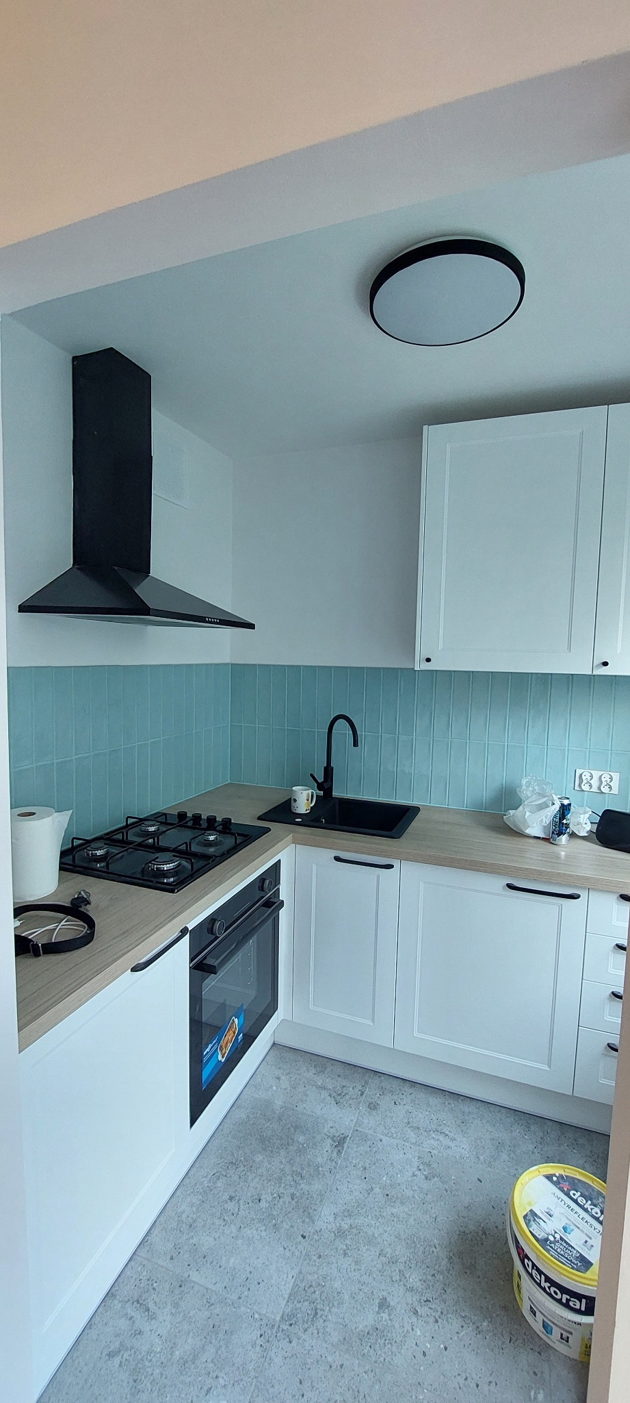 Nowoczesna, jasna kuchnia w Poznaniu z turkusowymi płytkami na ścianie, białymi szafkami z drewnianym blatem, czarnym zlewem i płytą grzewczą oraz widocznym wiaderkiem farby na podłodze.