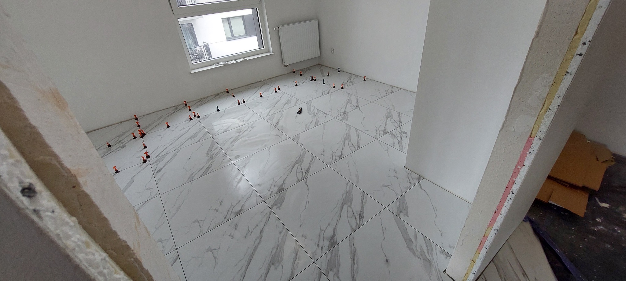 Układanie białych płytek podłogowych z szarym marmurkowym wzorem, z użyciem poziomujących klipsów i klinów w nowym, jasnym pomieszczeniu z oknem.