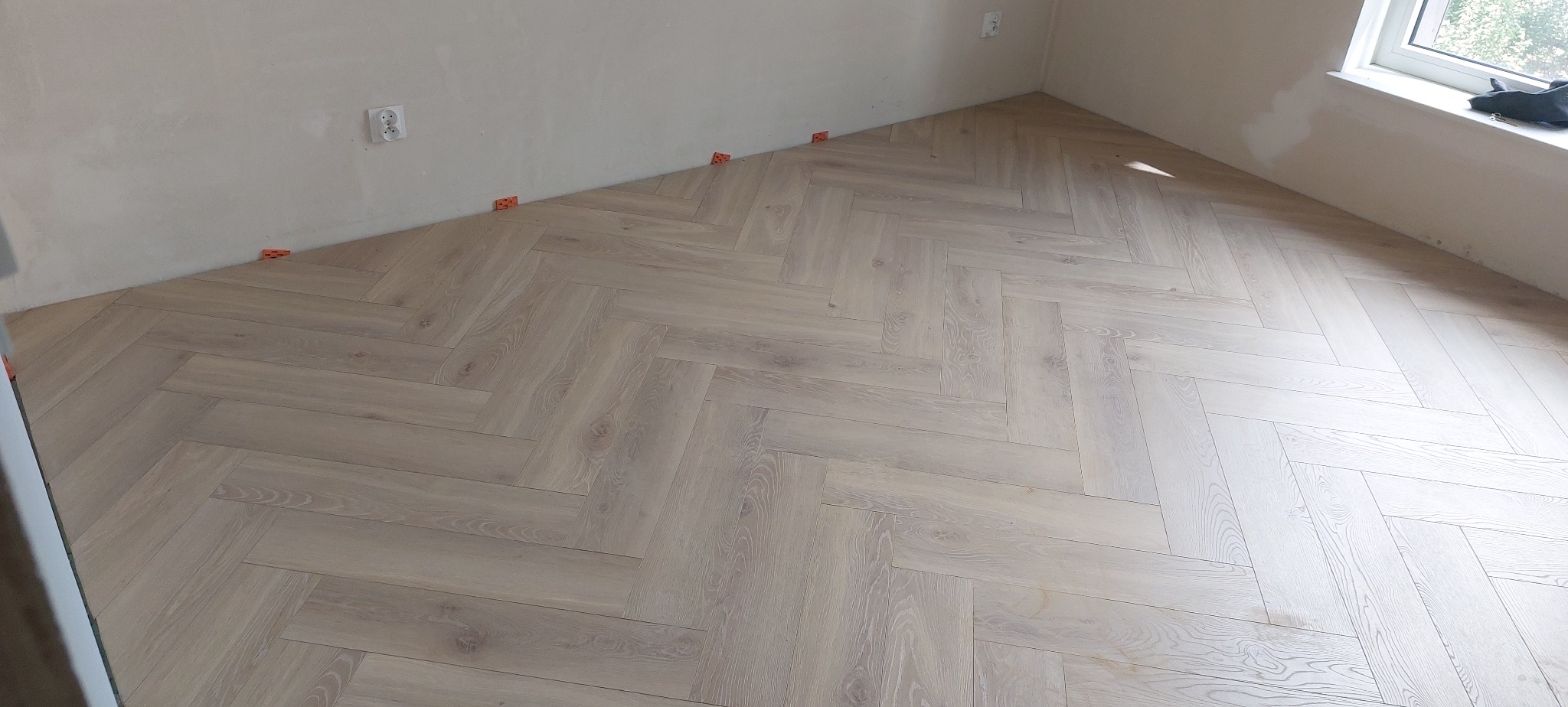 Jasne panele ułożone w jodełkę, widoczne pomarańczowe kliny dystansowe przy ścianie, świeżo wykończone ściany i okno.