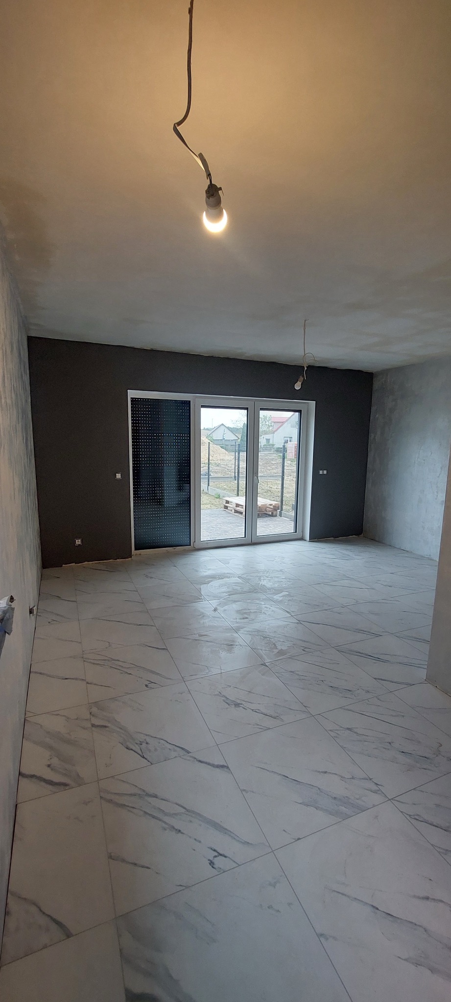 Wnętrze pokoju po remoncie z nową, białą podłogą z płytek imitujących marmur, szare ściany i surową żarówką zwisającą z sufitu.